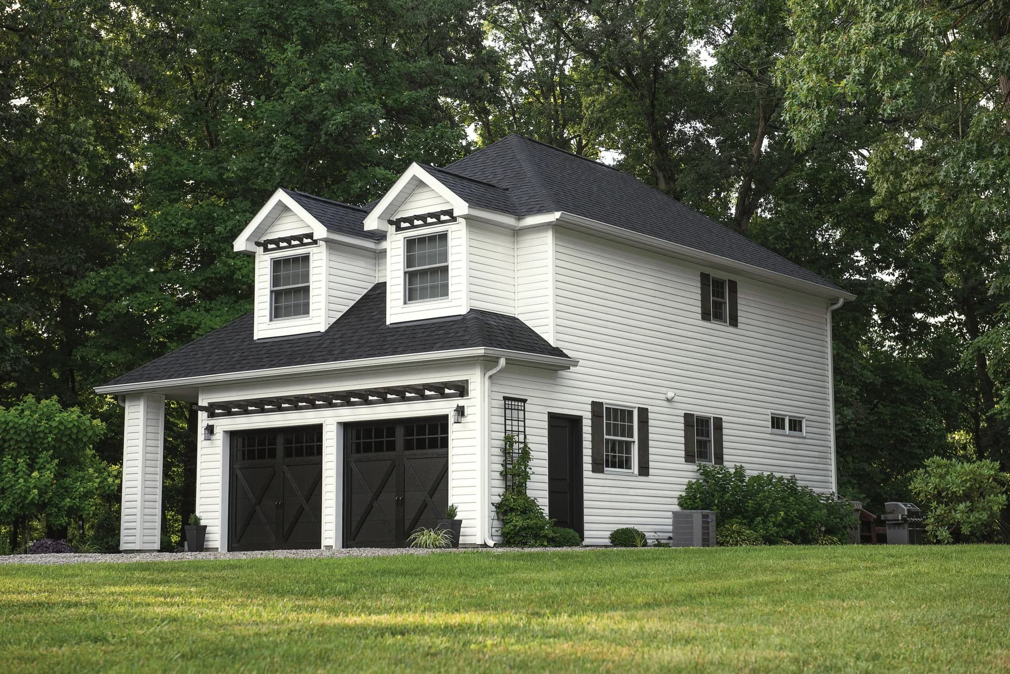 Two-story white carriage house with black shingled roof, dormer windows, and two dark carriage-style garage doors under a pergola