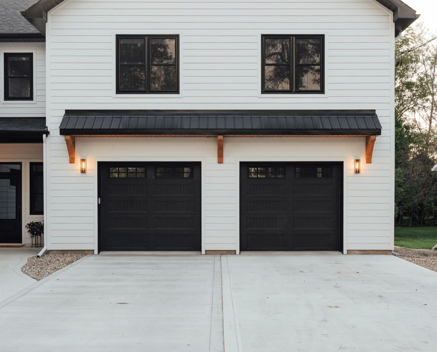 Black long-panel garage door under a metal awning on a white lap-sided home