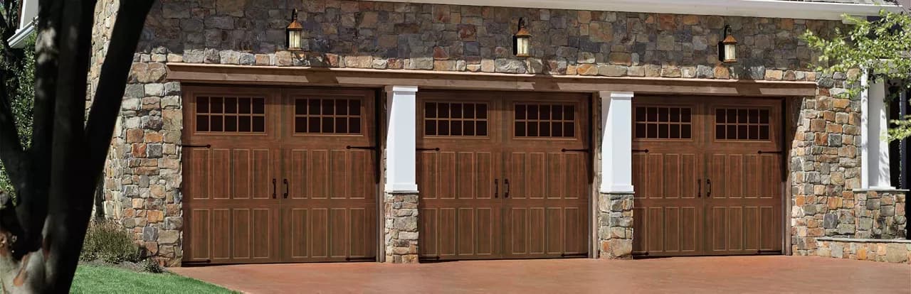 Three walnut-finish steel carriage-house garage doors with glazed top windows and decorative hardware on a stone-faced home