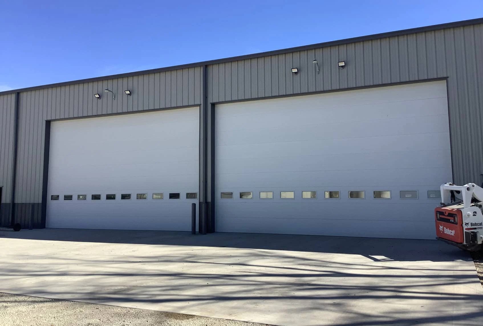 Two tall white insulated sectional steel doors with a row of vision panels, set into the grey metal wall of a shop building