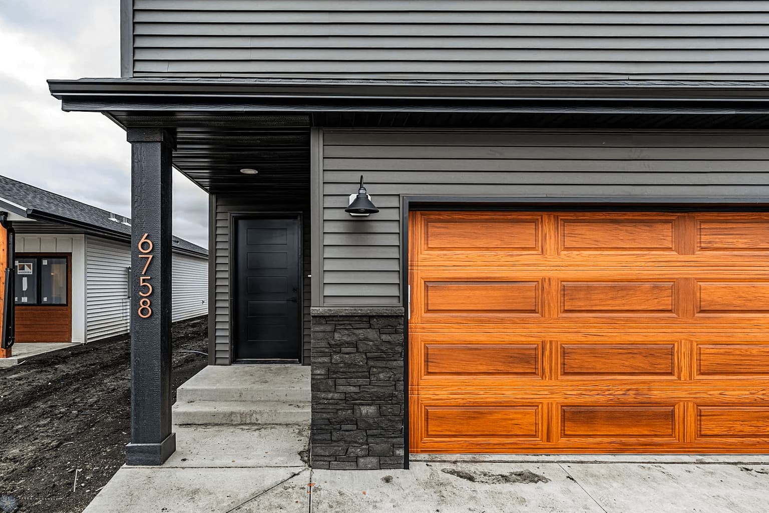 Cedar-stained raised-panel garage door on a gray-sided home with a stone-veneer column