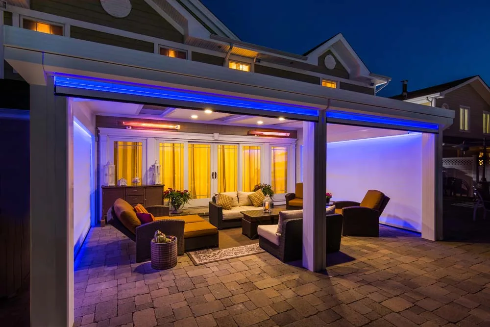 Covered backyard patio at night on a sided house, with screens lowered on both sides, a heater glowing, and wicker seating on pavers