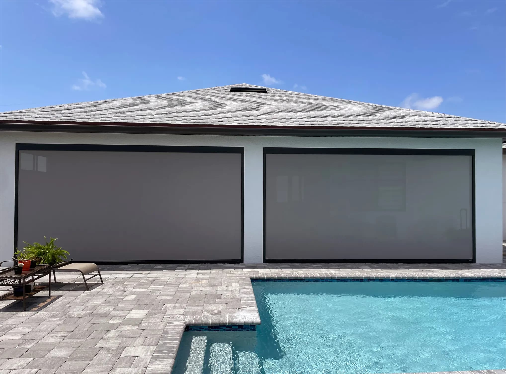 Two wide grey retractable screens lowered across the back of a white stucco house, with a paver patio and a pool in front