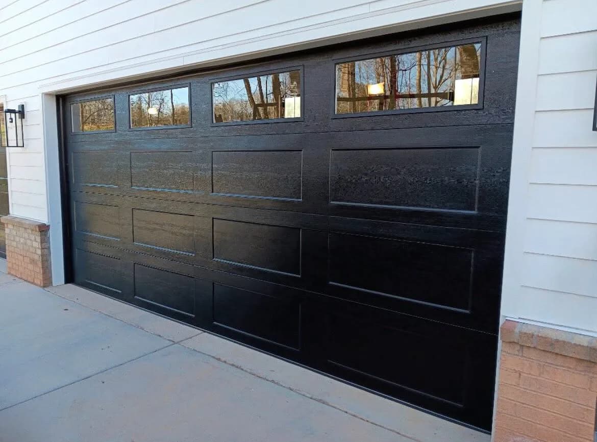 Black woodgrain long-panel garage door with a row of square top windows on a white lap-siding home with brick accents